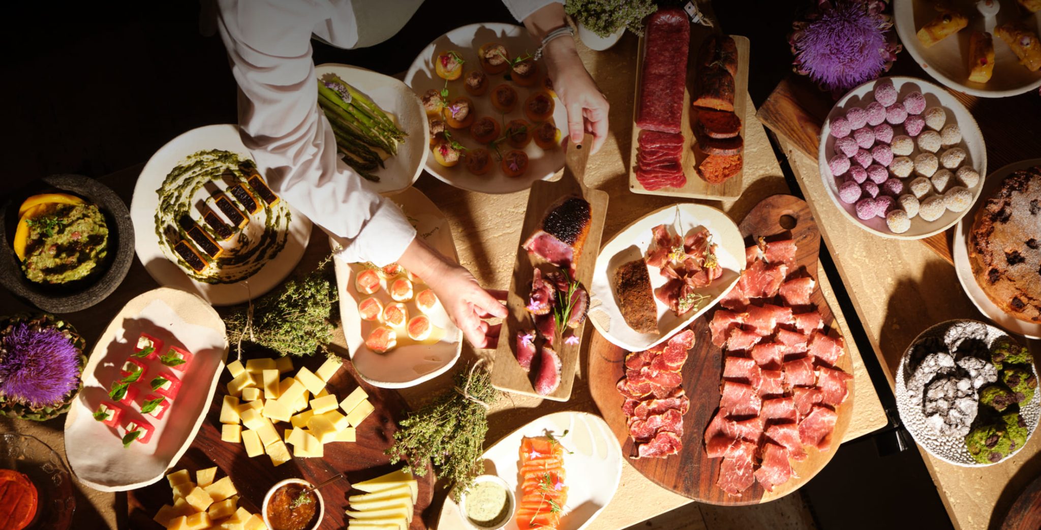 A long breakfast table seen from above, a cook plating a spread of cured meats, cheeses, fruit, cakes and herbs in warm low light