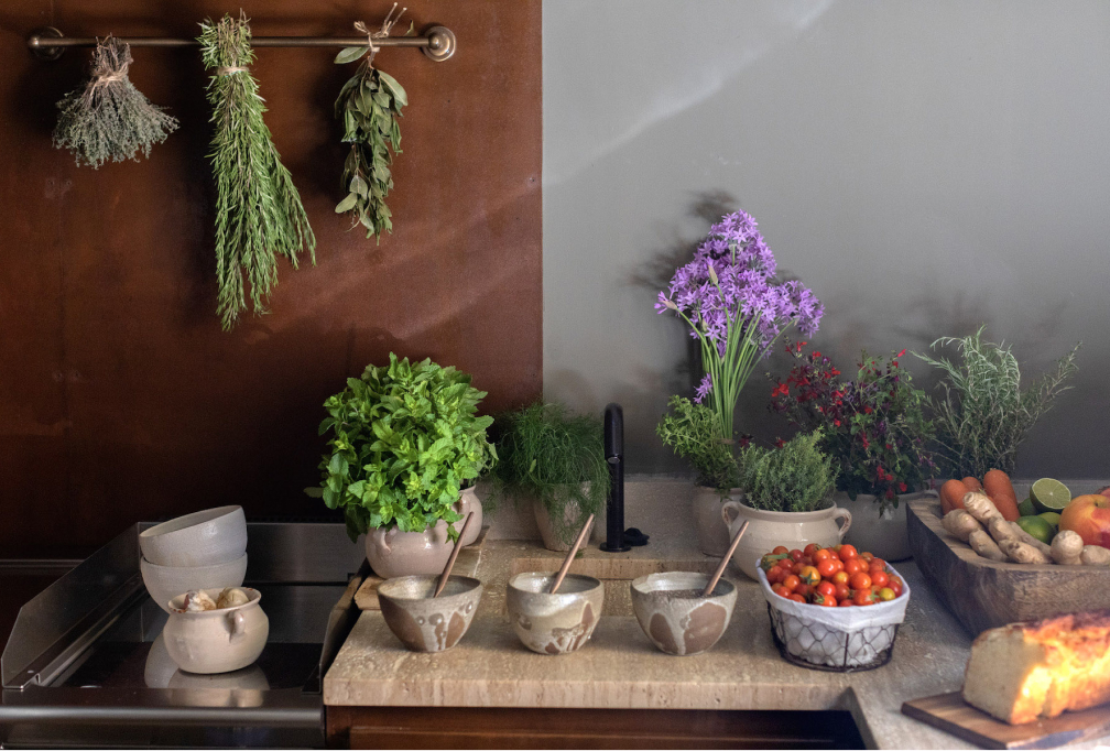 A kitchen prep counter with bunches of dried herbs hung on a rusted-steel wall, fresh basil and fennel in stoneware pots, cherry tomatoes and bread beside ceramic bowls
