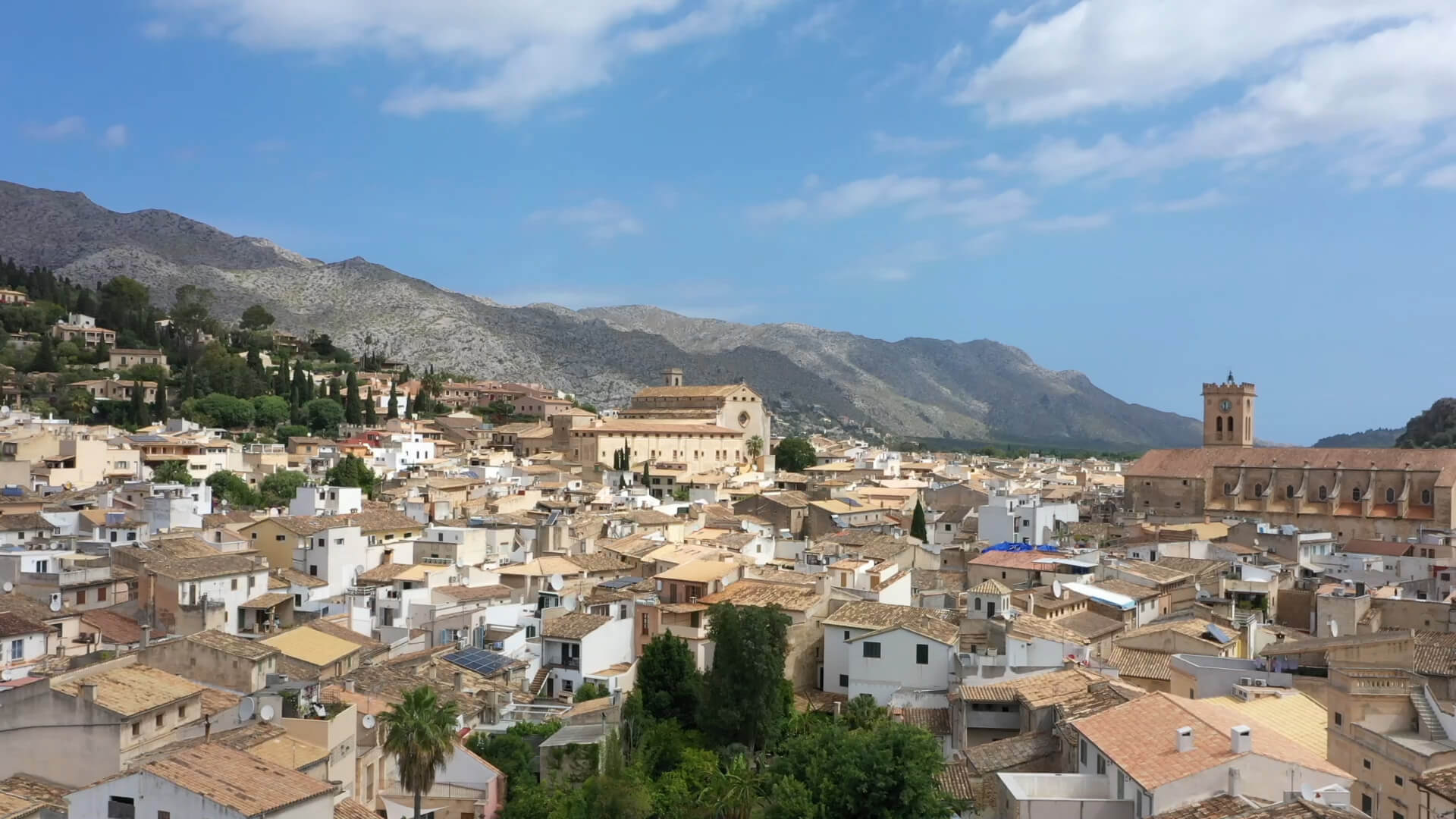 An aerial view over the terracotta rooftops of Pollença old town, the stone church and bell tower in the middle distance, the Serra de Tramuntana rising behind under a wide blue sky