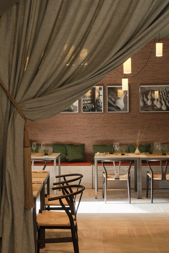 The From the Land dining room: a linen drape pulled back beside a brick wall hung with black-and-white photographs of Mallorcan pottery, wishbone chairs and laid tables under hanging lamps