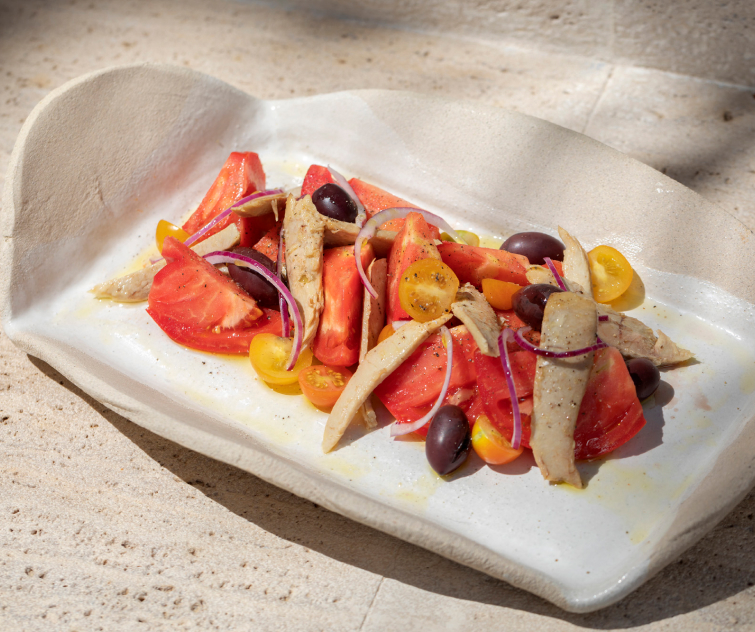 A plated dish of tomato and tuna from the From the Land kitchen, dressed simply with olives on ceramic in the sun