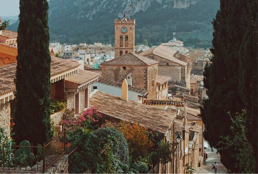 A view down through cypress trees and bougainvillea over the stone roofs of Pollença to the church bell tower and the mountains