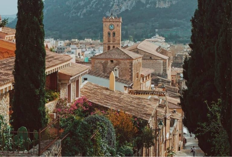 Pollença old town rooftops with the stone church tower in the middle distance and the Serra de Tramuntana rising behind under a wide blue sky