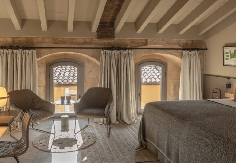 Stone-arch windows with white-painted beams overhead, two chairs in a pool of morning light in the Premium room at Can Aulí