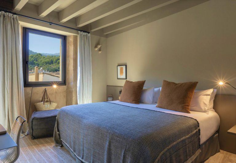 A room with a window framing the Tramuntana mountains directly, bed with terracotta cushions and island fabrics