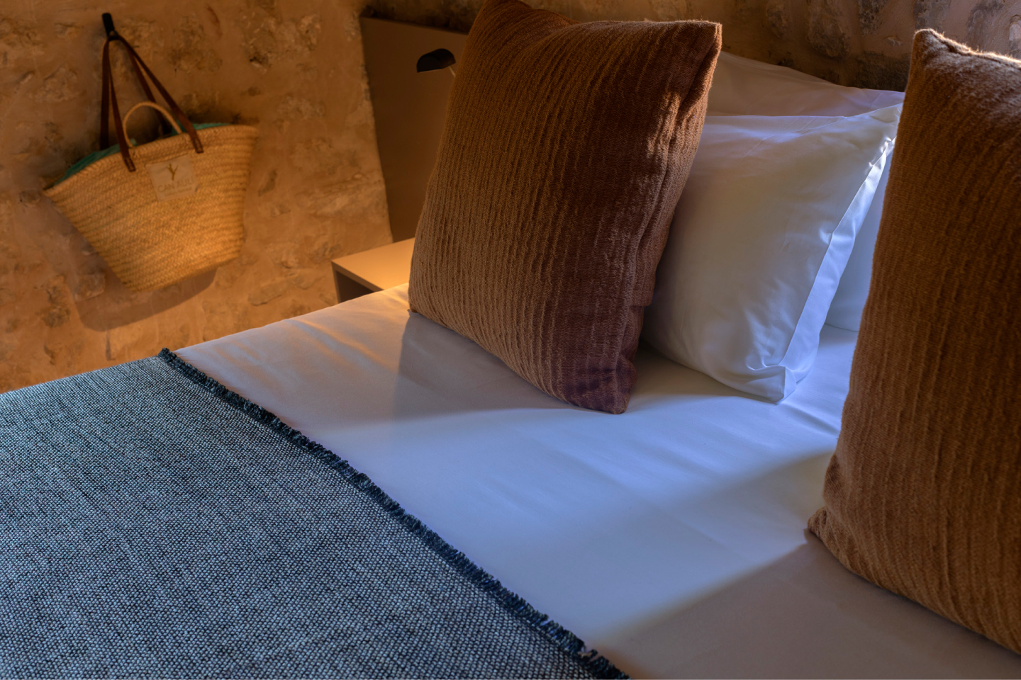 A made bed in warm low light against a bare marès stone wall, white linen with terracotta velvet cushions, an indigo woven throw folded across the foot, and a palm basket hung on the stone