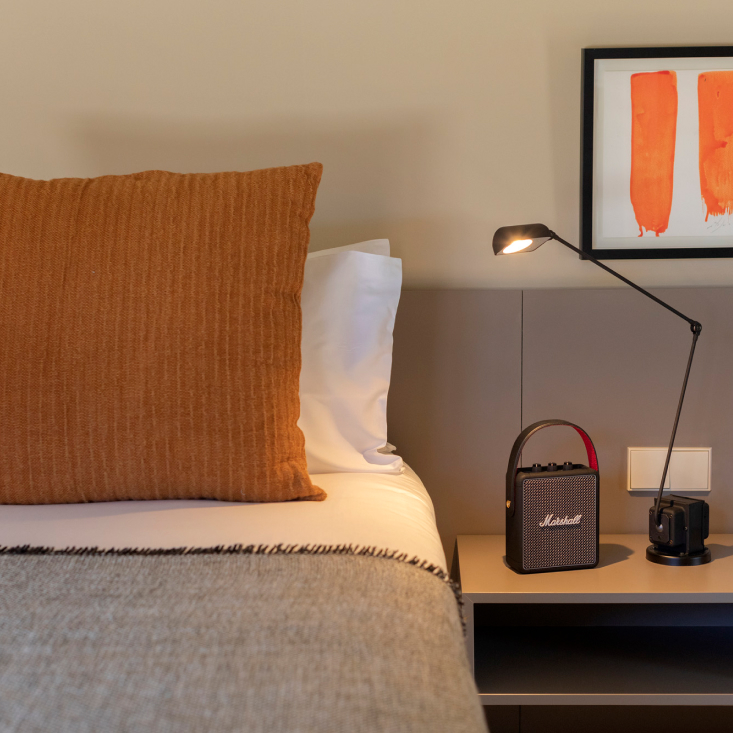 A corner of a stone room with white linen, a warm reading lamp and a woven palm basket against the bare wall
