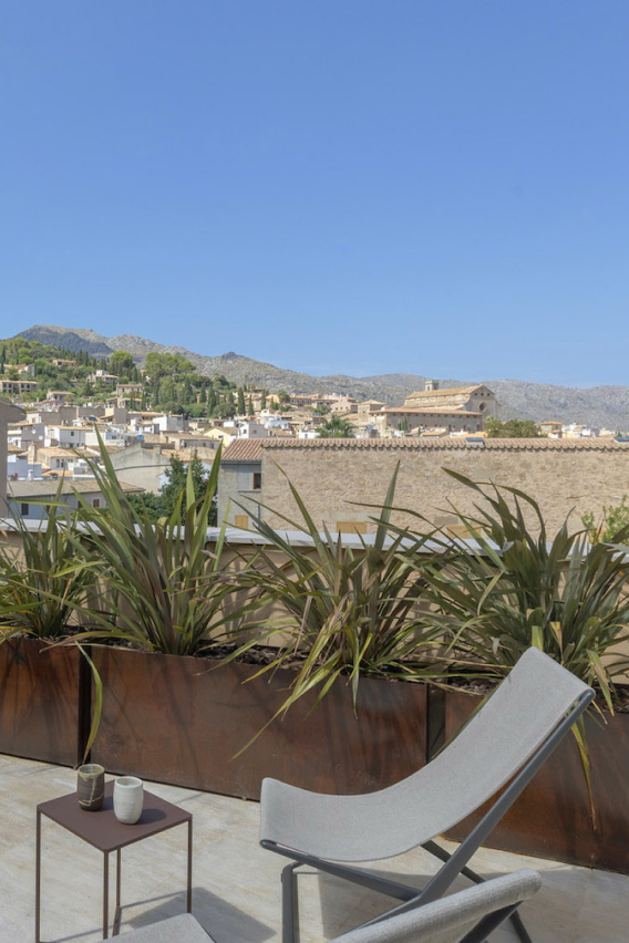 A private room terrace with a low chair and a small table, looking out over the stone rooftops of Pollença to the church and the Serra de Tramuntana under a clear sky